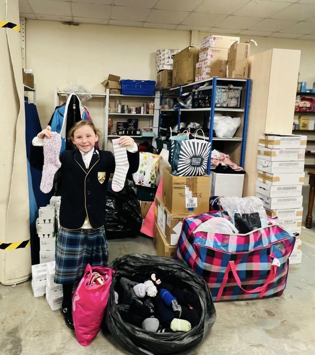 Elsie in her school uniform holding up two pairs of socks, with bags full of donated socks at her feet